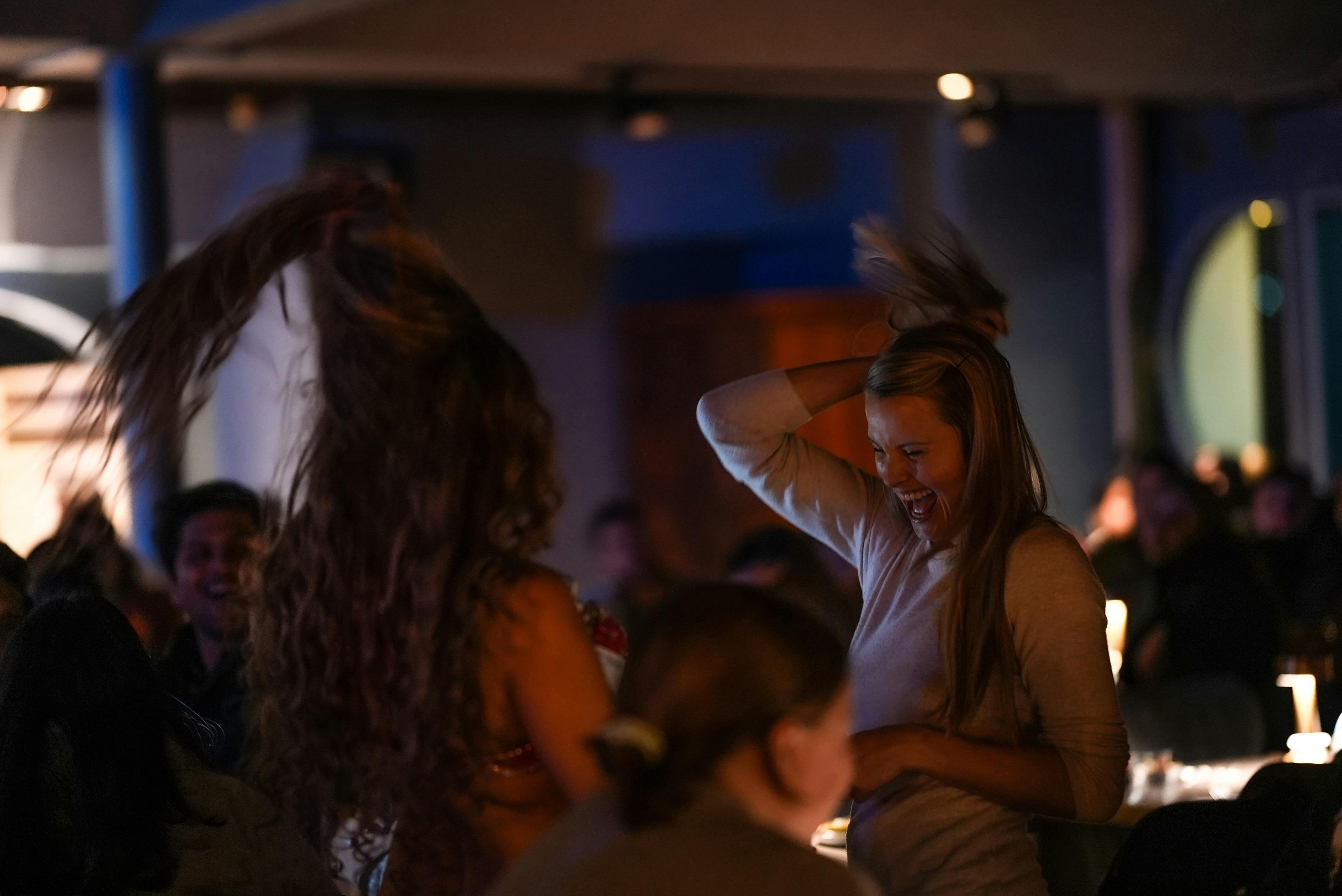 長い髪の女性が笑顔で屋内で踊っていて、薄暗く照らされた群衆に囲まれています。