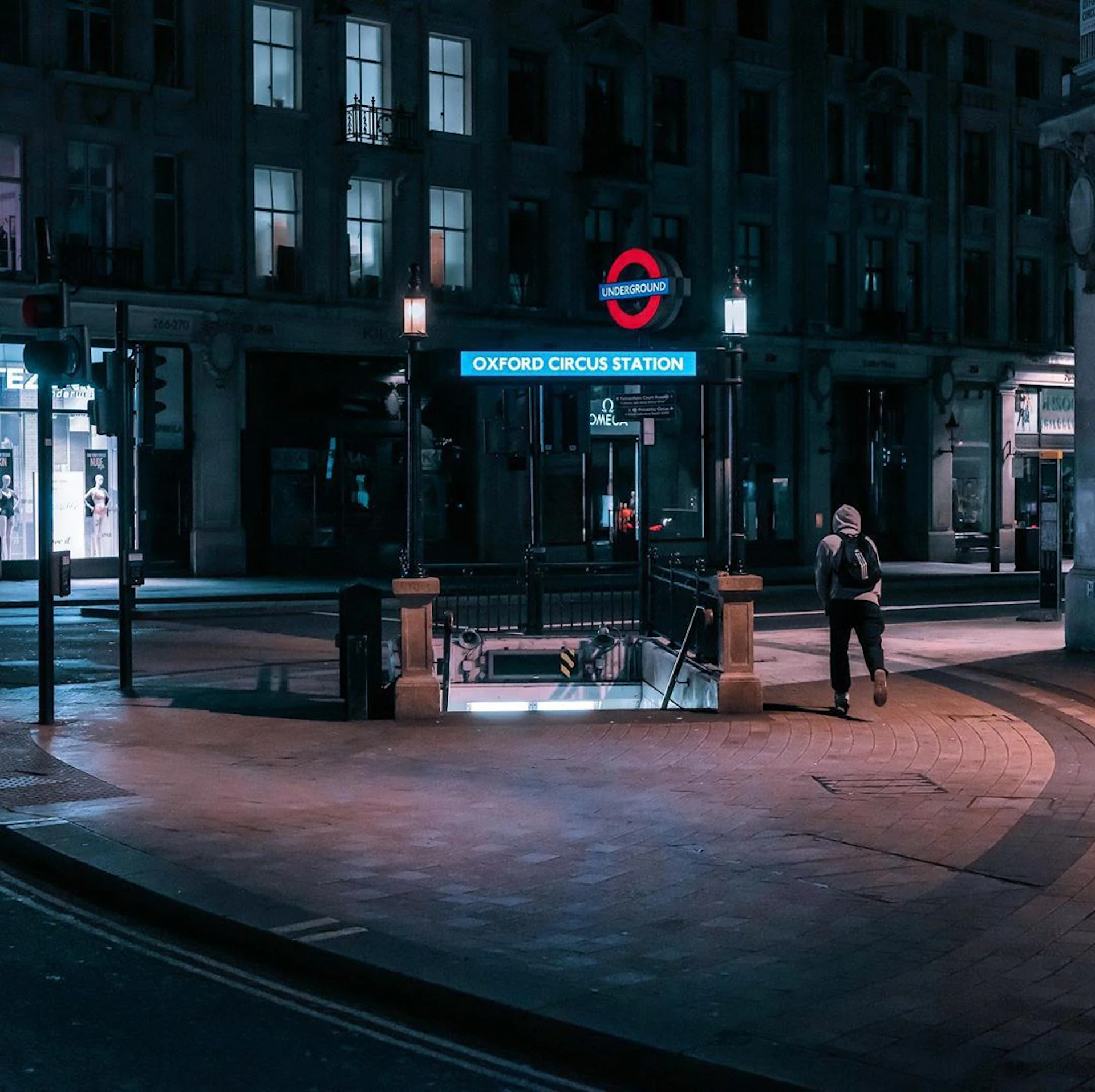 A person in a hoodie walks by the Oxford Circus Station entrance, illuminated in an empty, quiet street at night.