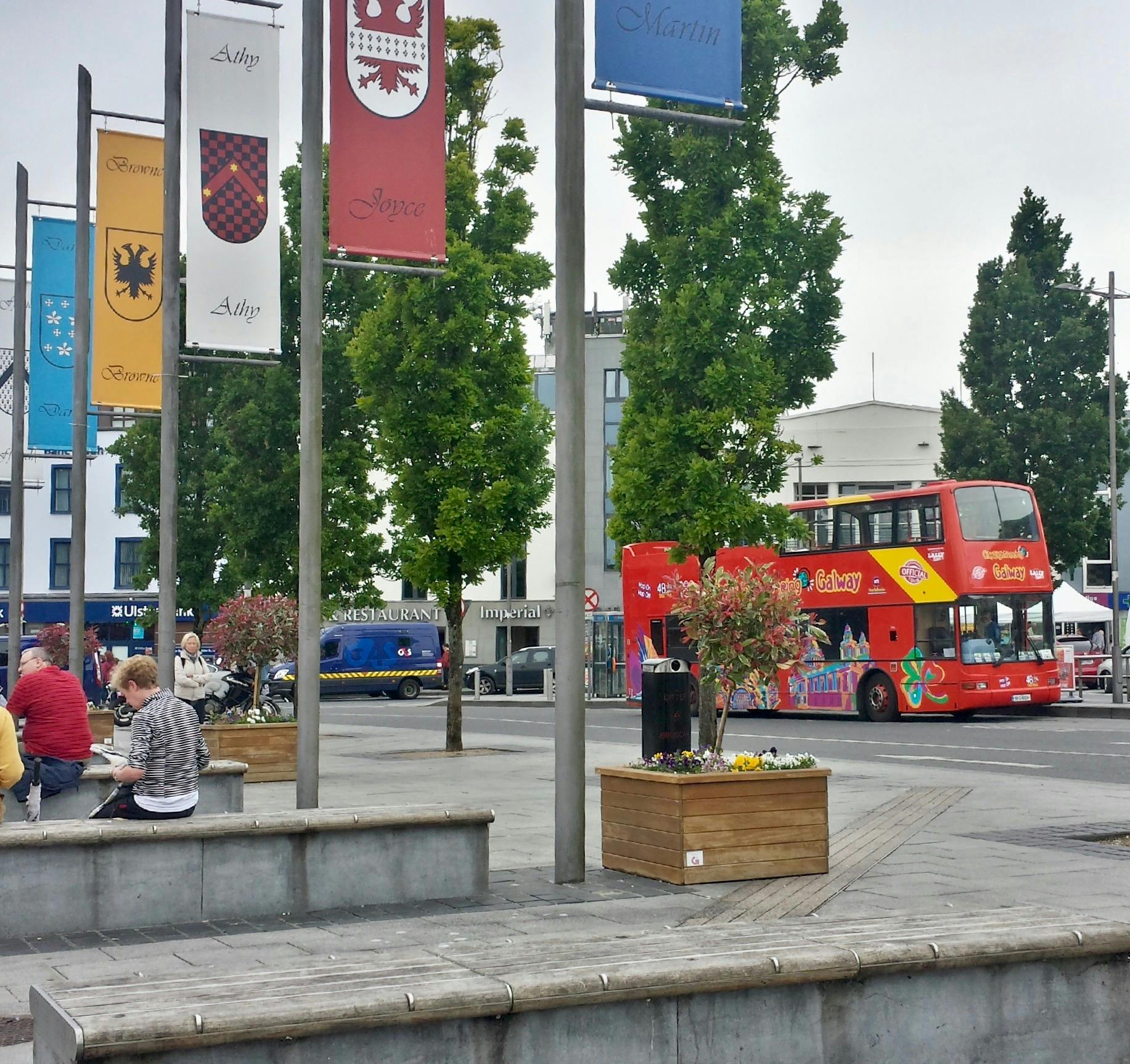 Urban plaza with flagpoles, benches, planters, and people sitting. Red double-decker tour bus and blue van on adjacent street.