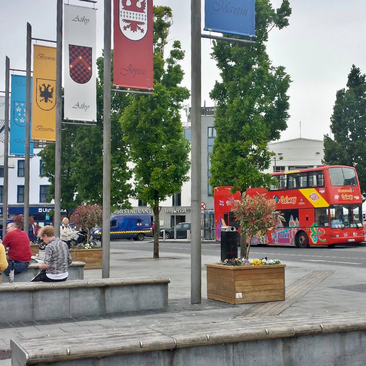 Urban plaza with flagpoles, benches, planters, and people sitting. Red double-decker tour bus and blue van on adjacent street.