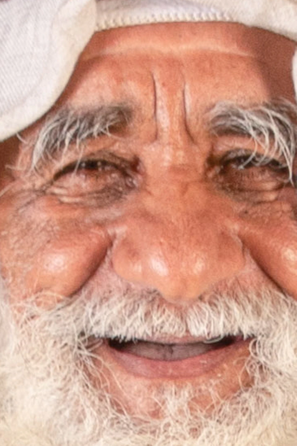 An elderly man with a long white beard, wearing traditional attire, holds fishing net repair tools against a beige background with a draped net.