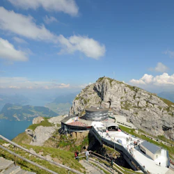 Människor som går på stengångar och plattformar på ett berg med naturskön utsikt över en sjö och dalar under en blå himmel.