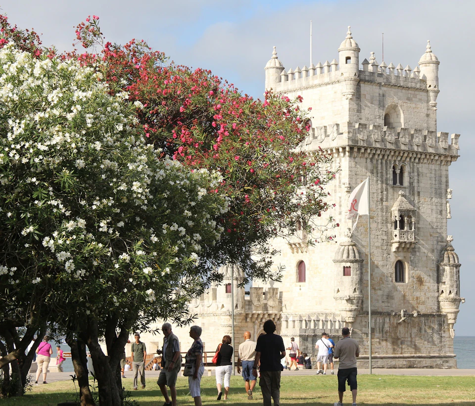 People walking beside a large, historic stone tower with uniquely shaped parapets and surrounded by lush green trees with pink and white flowers.