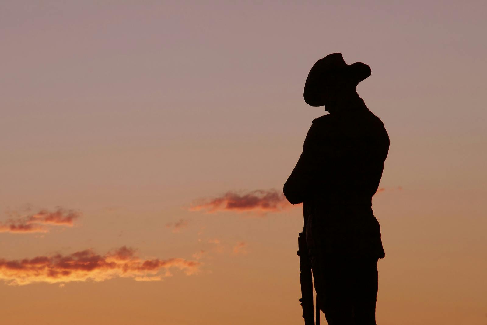 Silueta de una estatua de soldado sosteniendo un fusil al atardecer, con un cielo rosa claro y naranja de fondo.