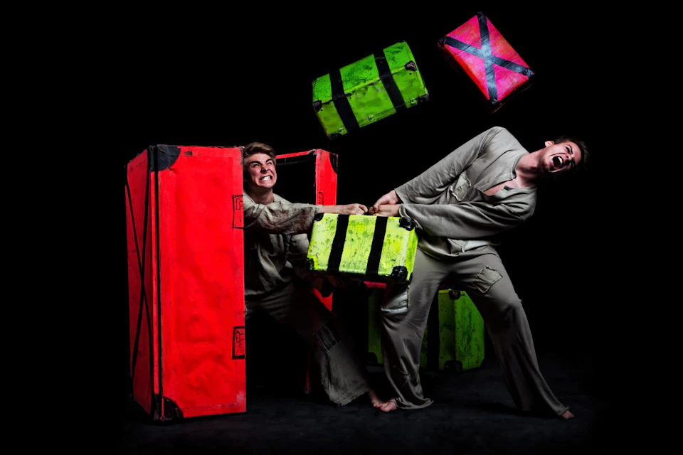 Two people pulling on bright colored crates, one in red and the other in green, against a dark background.