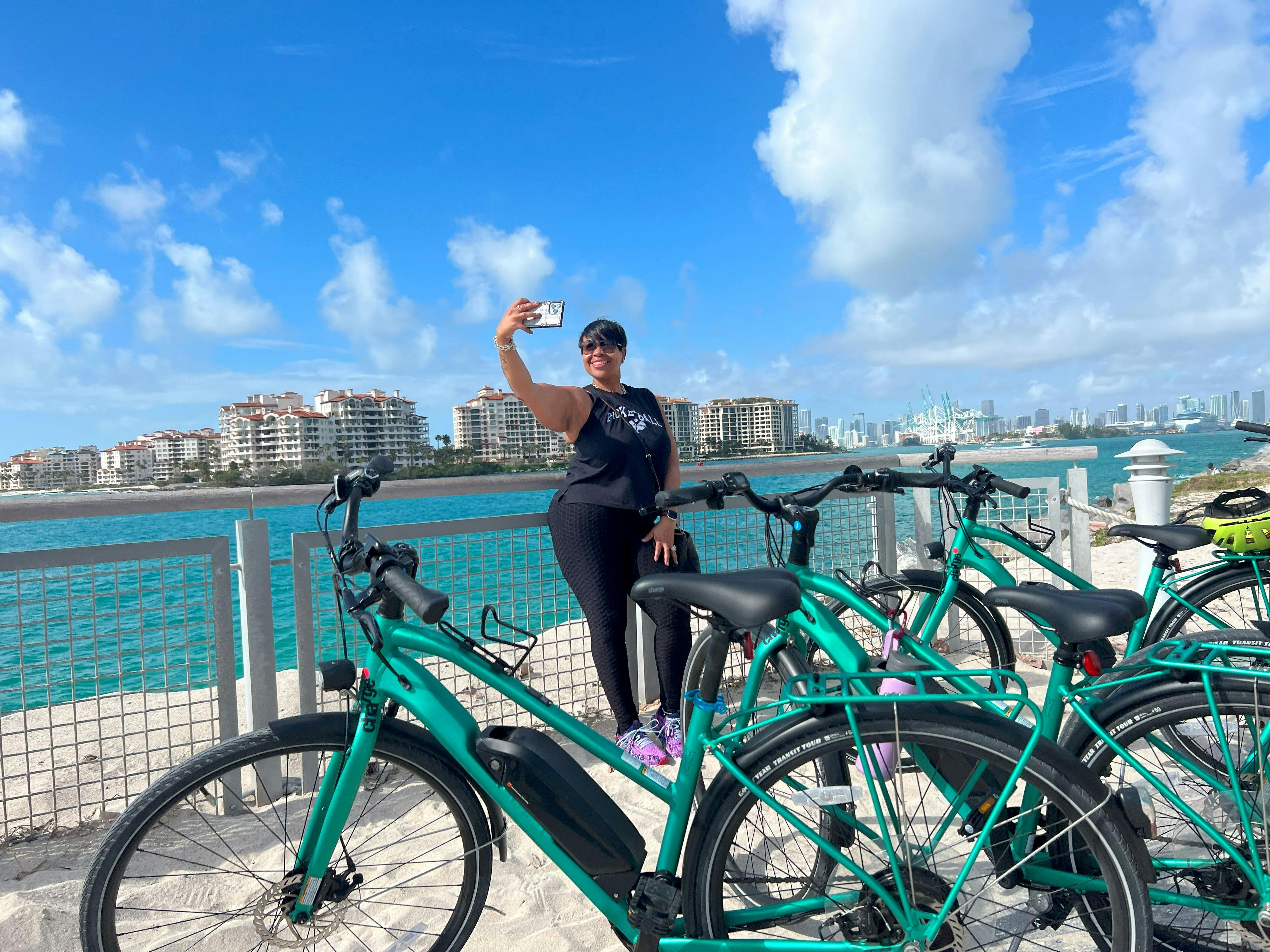 Člověk si udělá selfie poblíž tyrkysových kol na nábřeží s městskými budovami a částečně zataženou oblohou v pozadí.