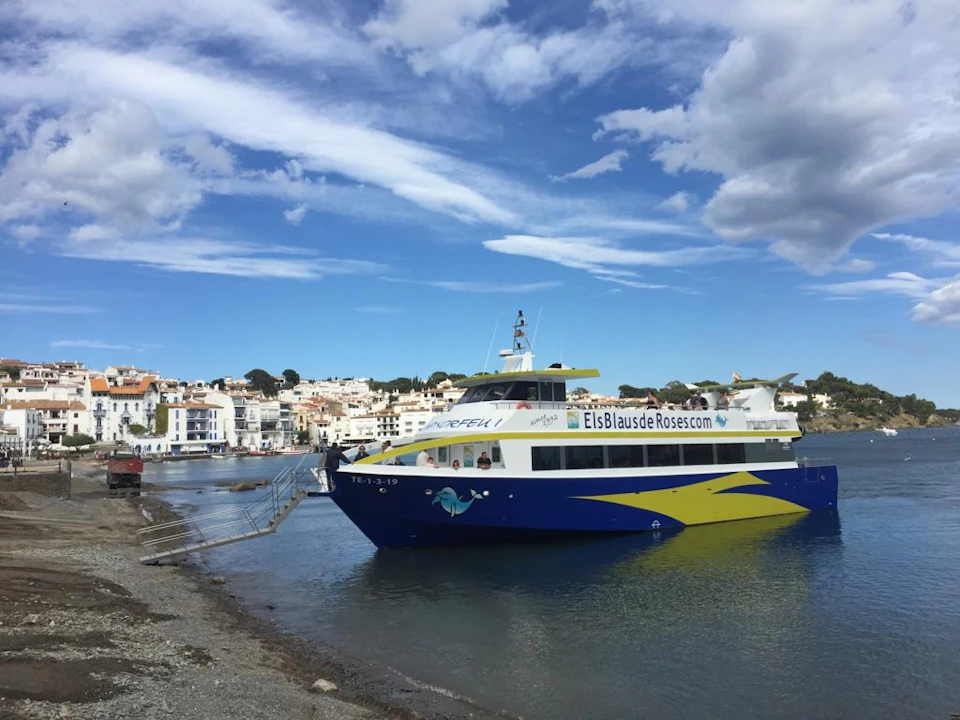 A blue and yellow boat docked near a coastal town with white buildings under a blue sky with scattered clouds.