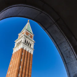 Klocktorn i tegel med en grön spiral sedd genom en stenbåge, mot en klarblå himmel.
