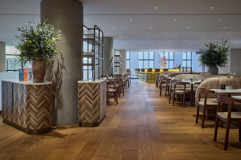 A modern restaurant interior with wooden floors, tables, chairs, and a bright bar area in the background.