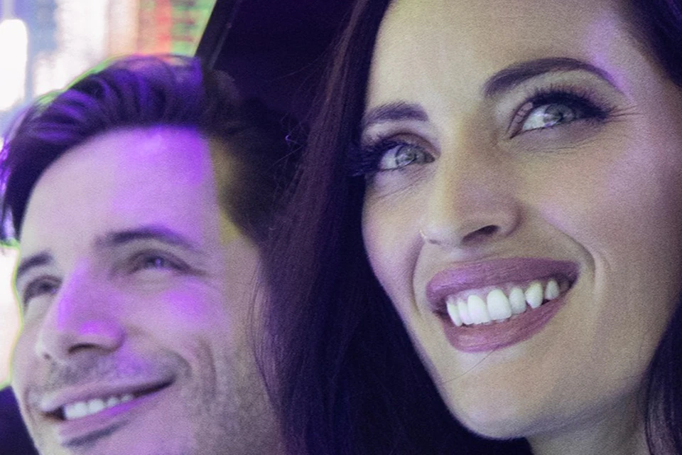 Two people smiling while standing next to a reflective and colorful light installation.