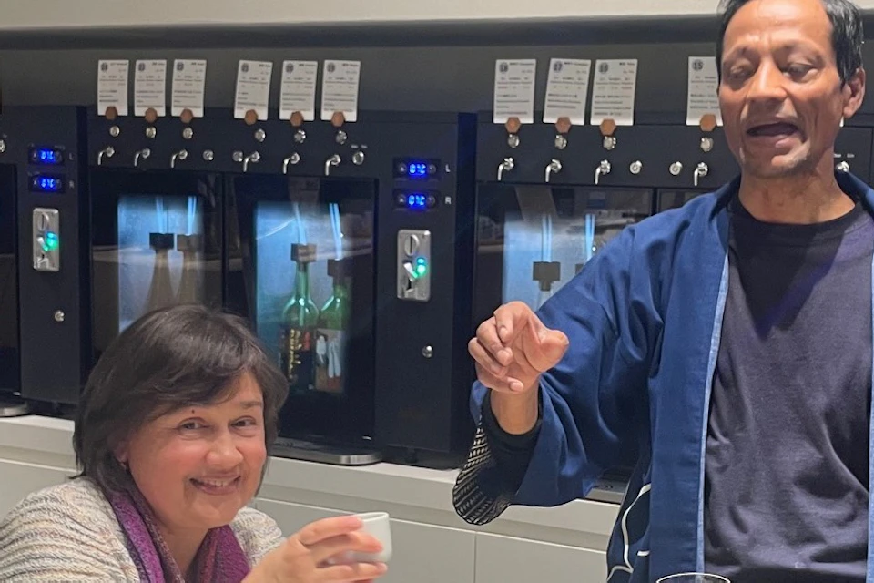 Four smiling people raising glasses at a table with a sake tasting setup. Background shows sake dispensers and display bottles.
