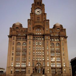 En stor, utsmyckad byggnad med ett framträdande klocktorn och många fönster, toppad med en fågelskulptur, under en klar himmel.