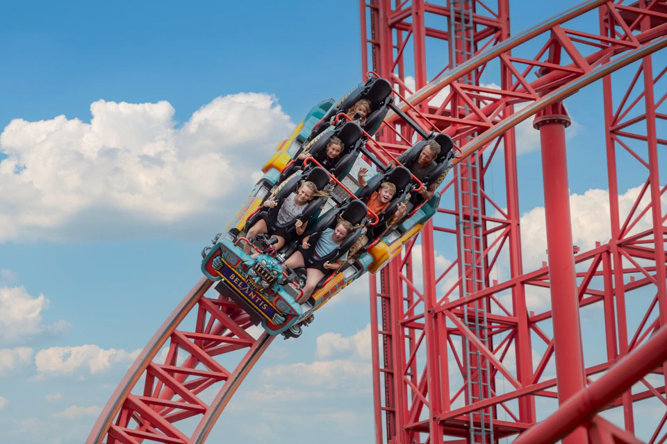 A roller coaster with passengers is descending a steep red track under a blue sky with scattered clouds.