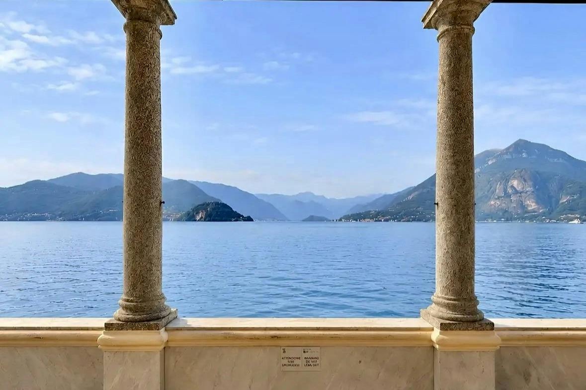 A serene lake view framed by two stone columns with distant mountains and a clear blue sky in the background.