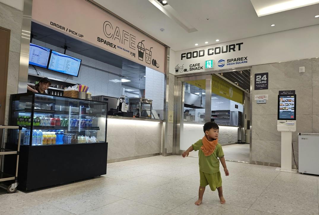 A child in green clothing stands barefoot in a food court area with signage and a digital kiosk visible.