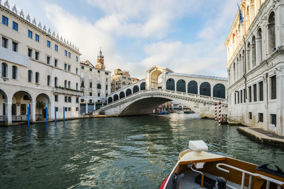 Stone bridge with arches spans over a canal, surrounded by historic buildings. A boat's bow is visible in the foreground.