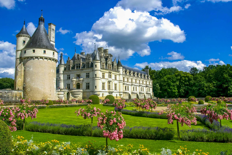 A grand castle with turrets and manicured gardens, featuring pink flowering trees and green lawns under a blue sky with clouds.