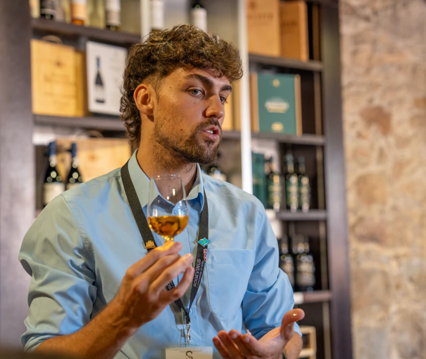 A person with a blue shirt and lanyard holds a glass of wine, speaking in a room lined with wine bottles and boxes.