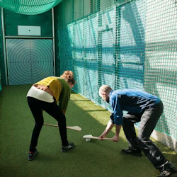 Två personer spelar golvhockey i ett slutet nätområde, med fokus på ett övningsmål i slutet.