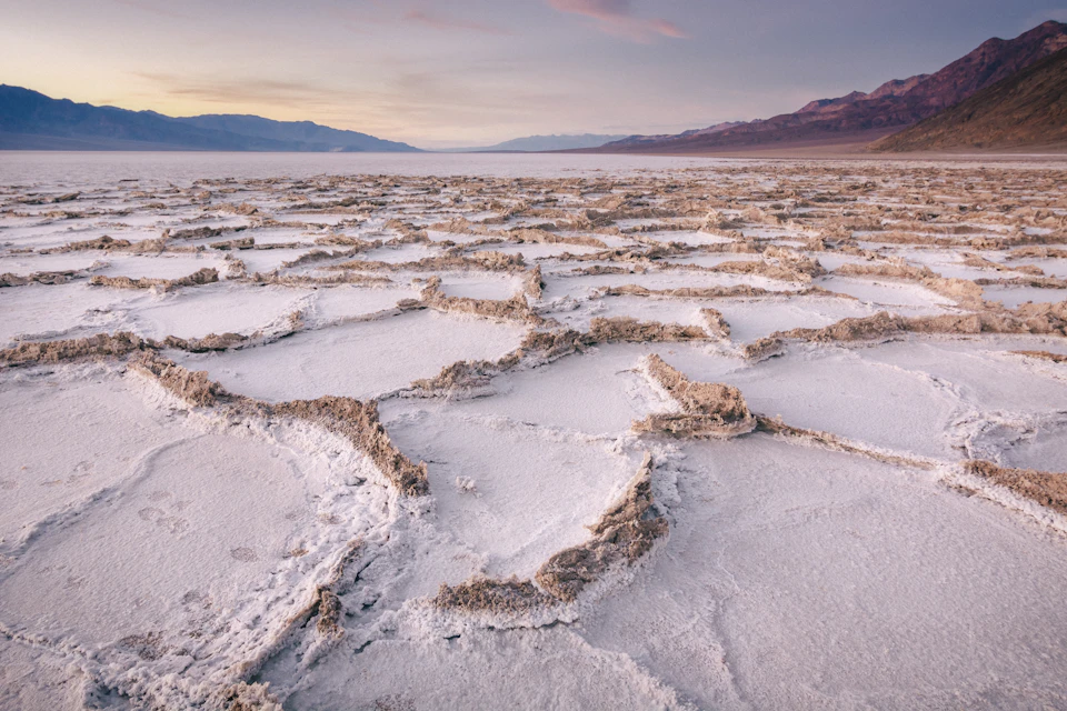 Salt flats with hexagonal patterns stretch towards the horizon, with mountains and a pastel-colored sky in the background.