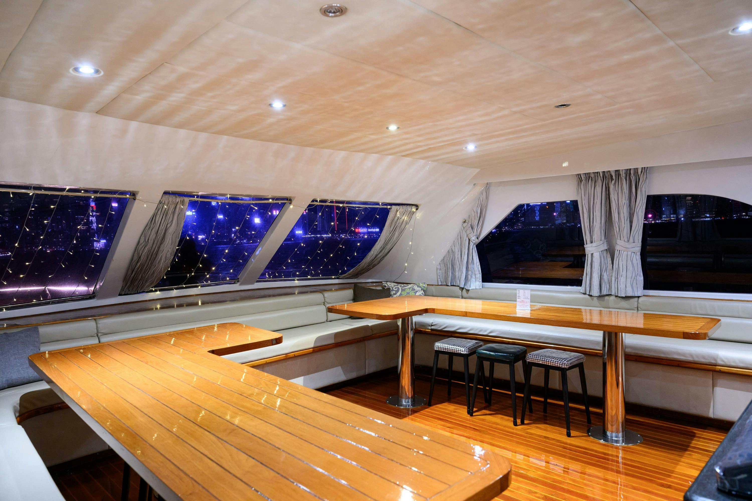 An interior of a boat with wooden tables, metal stools, and large windows decorated with string lights, showing city lights outside.