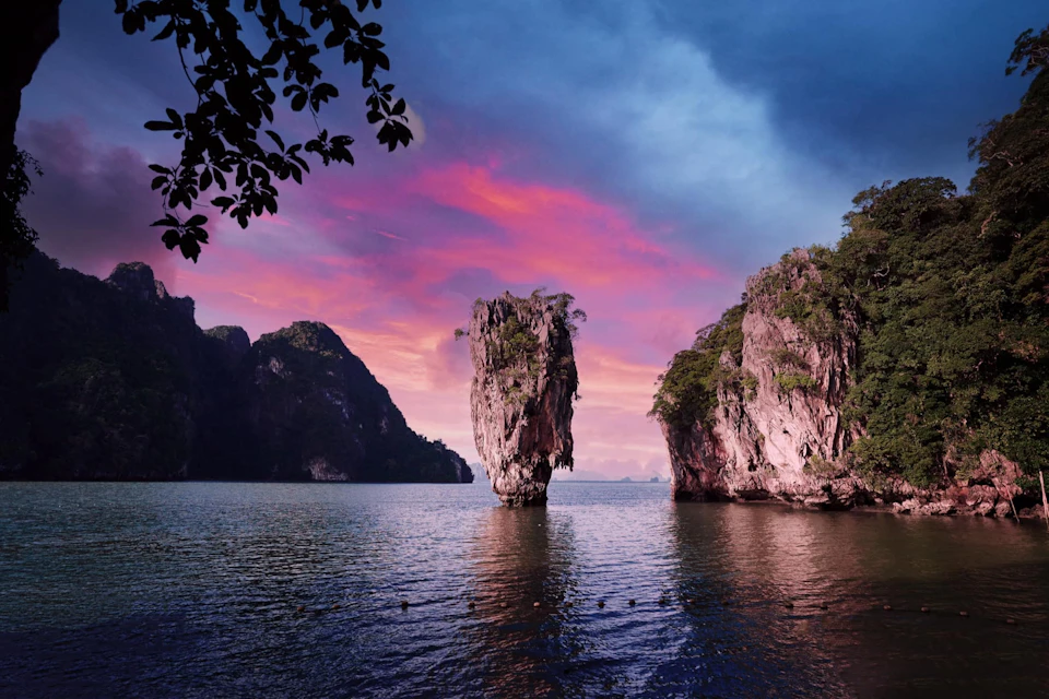 A serene seascape with a tall, vertical rock formation in the water against a vibrant purple and pink sunset sky, surrounded by cliffs.