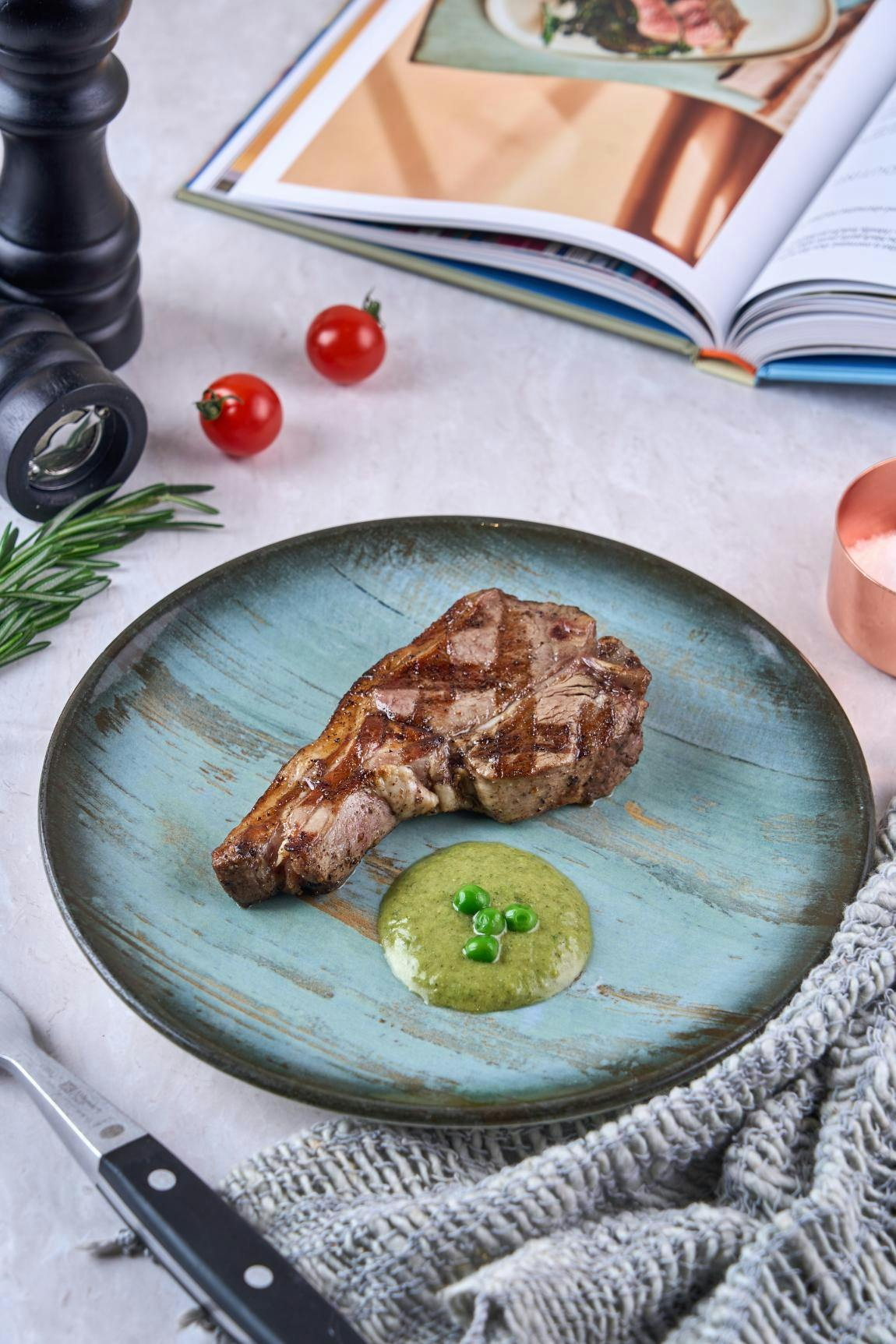 Grilled meat on a blue plate with green puree and peas, garnished with cherry tomatoes and rosemary on the side.