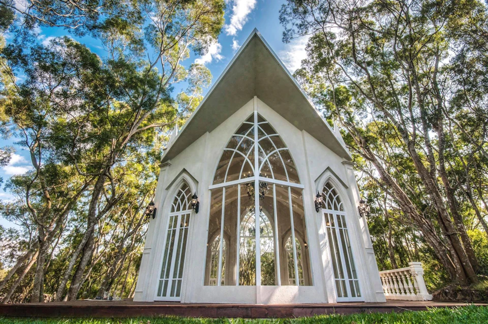 A small white chapel with large arched windows surrounded by tall trees under a blue sky with scattered clouds.