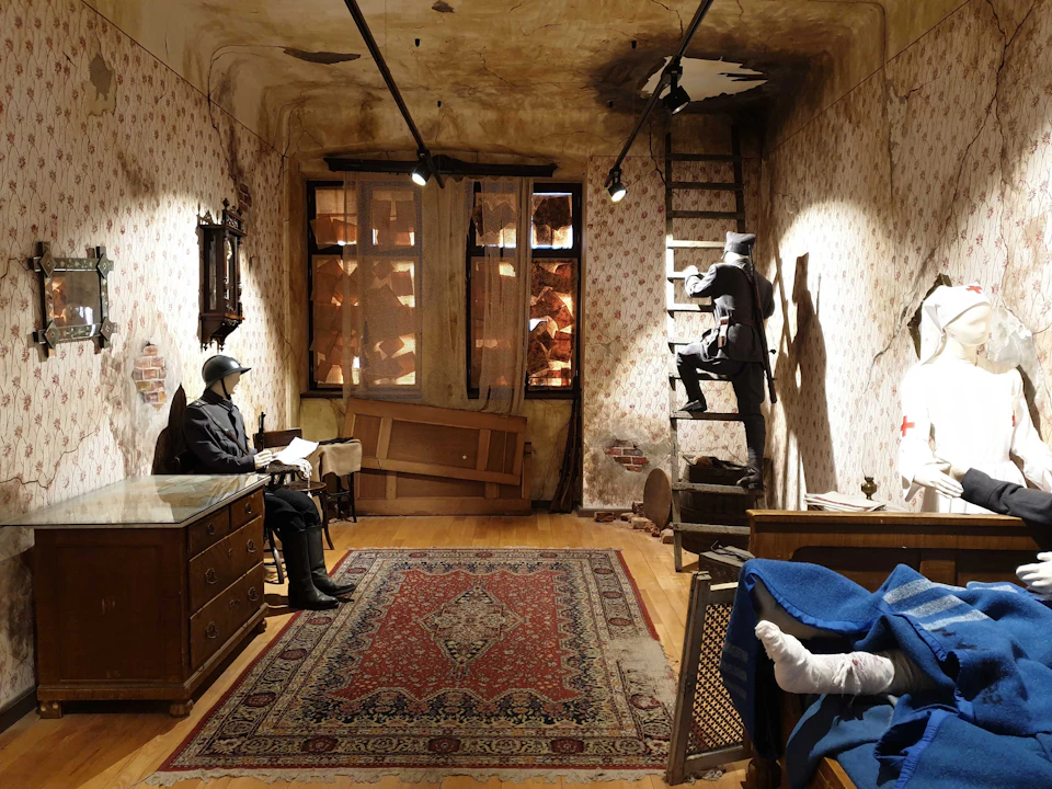 A room with damaged wallpaper, featuring a soldier mannequin reading at a desk and another climbing a ladder. A person is lying in bed.