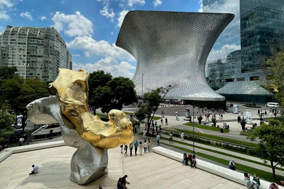 People walking around a modern plaza featuring a large, unique silver building and a gold and silver abstract sculpture.