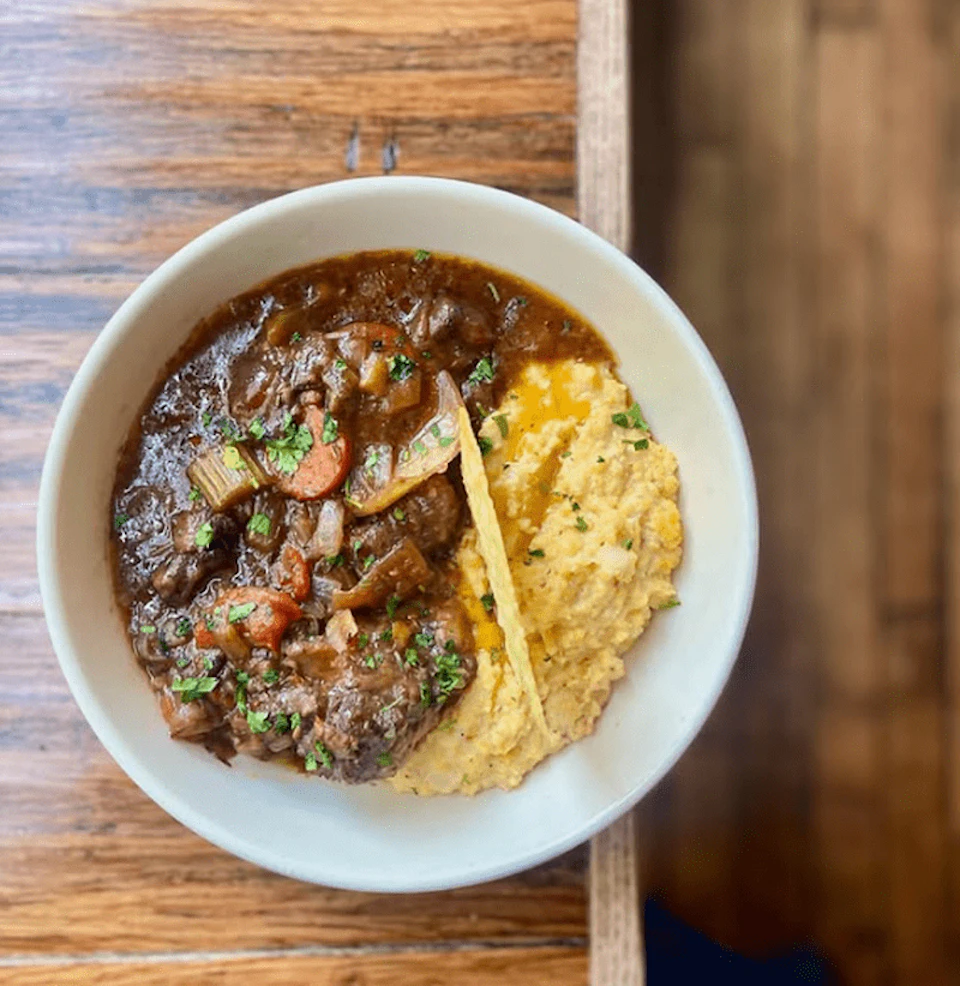 A bowl of beef stew with vegetables and polenta on a wooden table.