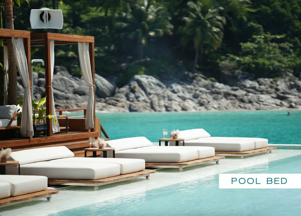 Lounge area with white sunbeds and small tables beside a turquoise pool, set against a backdrop of rocks and lush greenery.