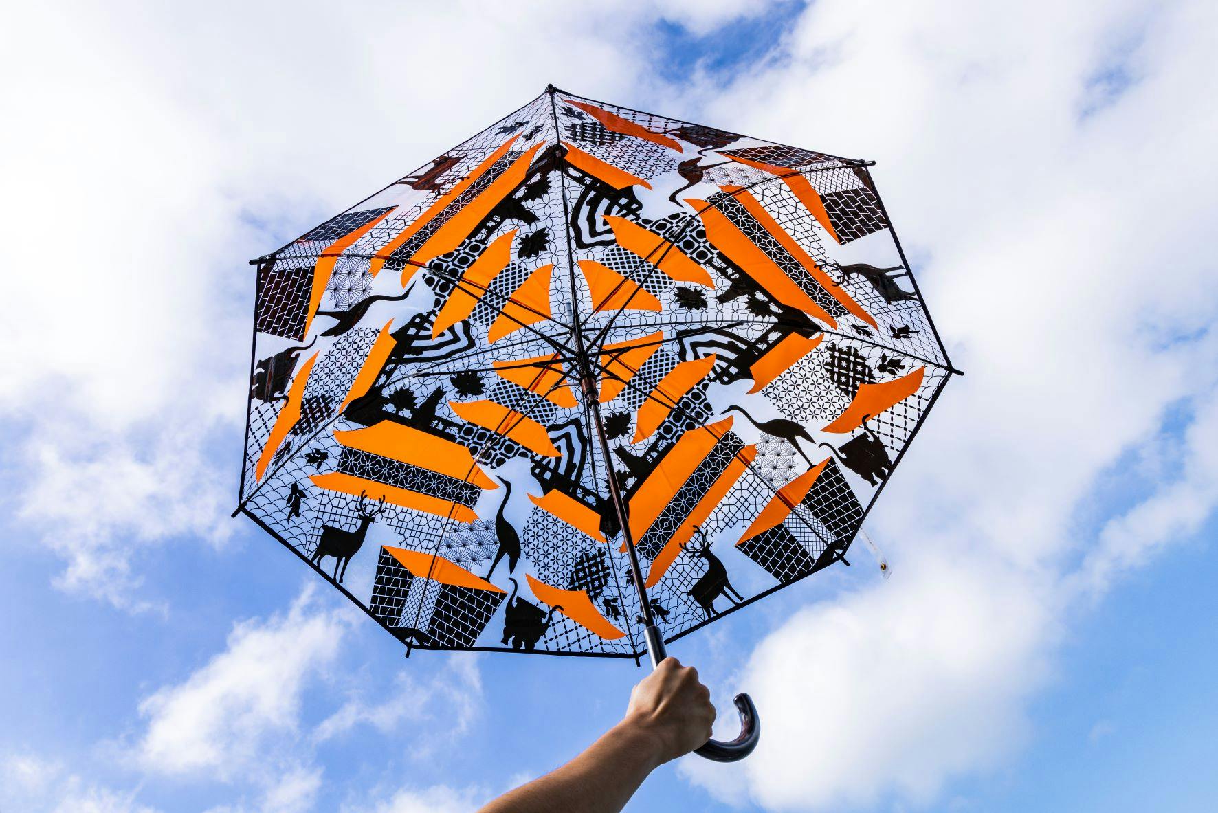 A hand holds a colorful umbrella with orange, black, and white patterns, set against a partly cloudy blue sky.
