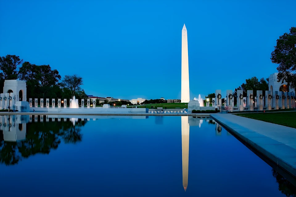 The Washington Monument reflected in a calm pool of water, with surrounding structures and a clear evening sky.