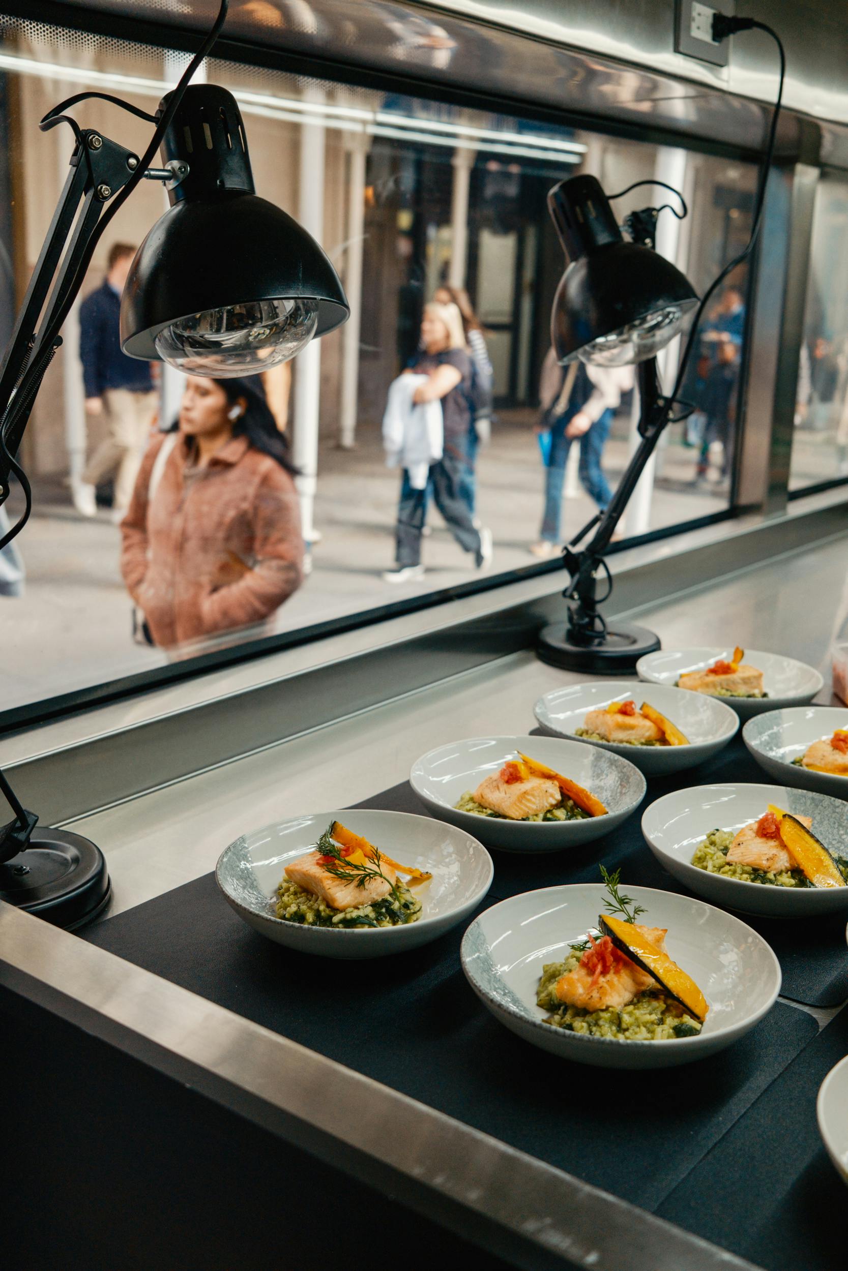 Schalen salade met zalm op een toonbank, verlicht door lampen, met mensen die buiten een raam lopen in een stedelijke omgeving.