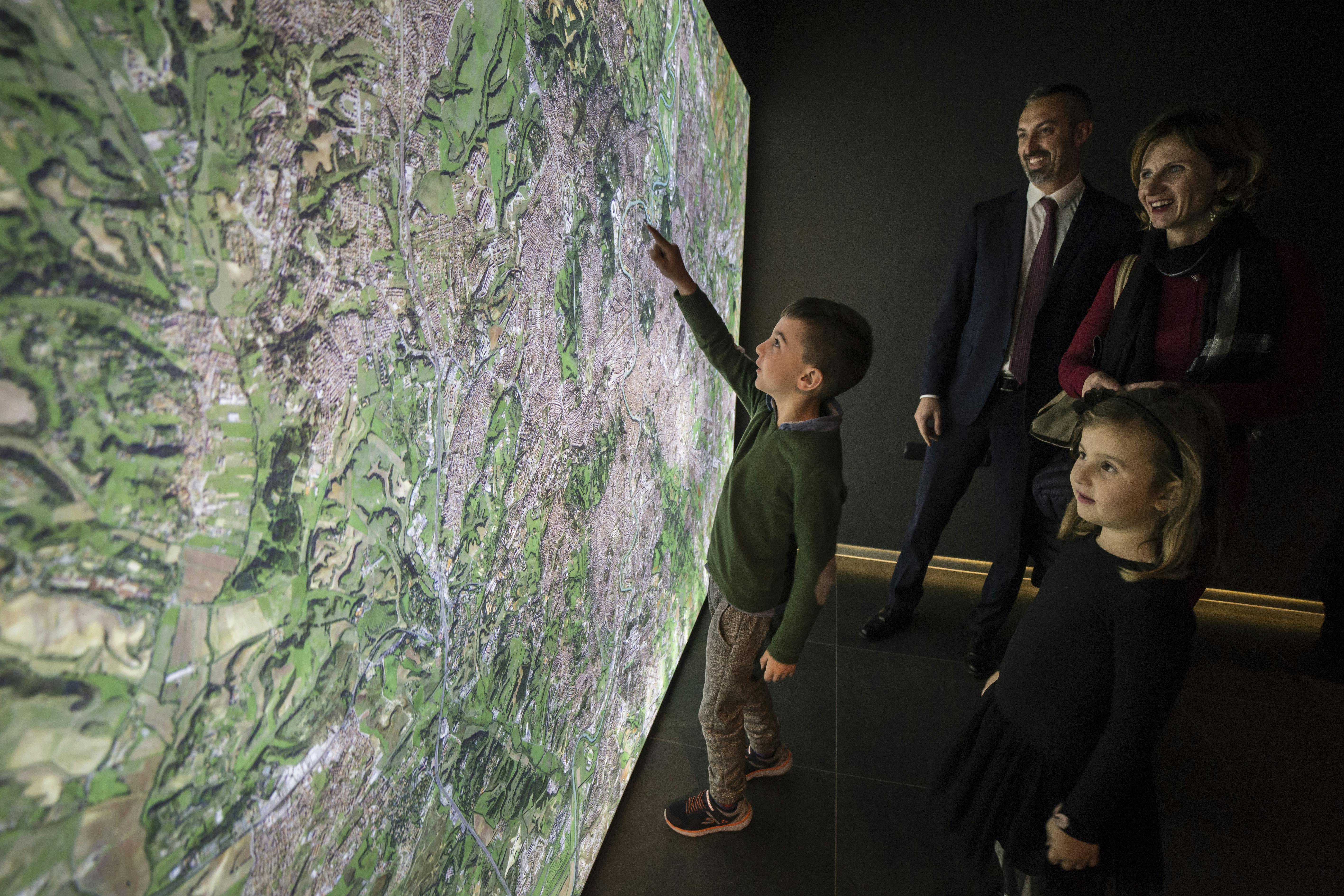 한 소년이 벽에 비춰진 커다란 지도를 가리키고 두 명의 성인과 한 소녀가 어두운 방 안에서 미소를 짓고 있습니다.