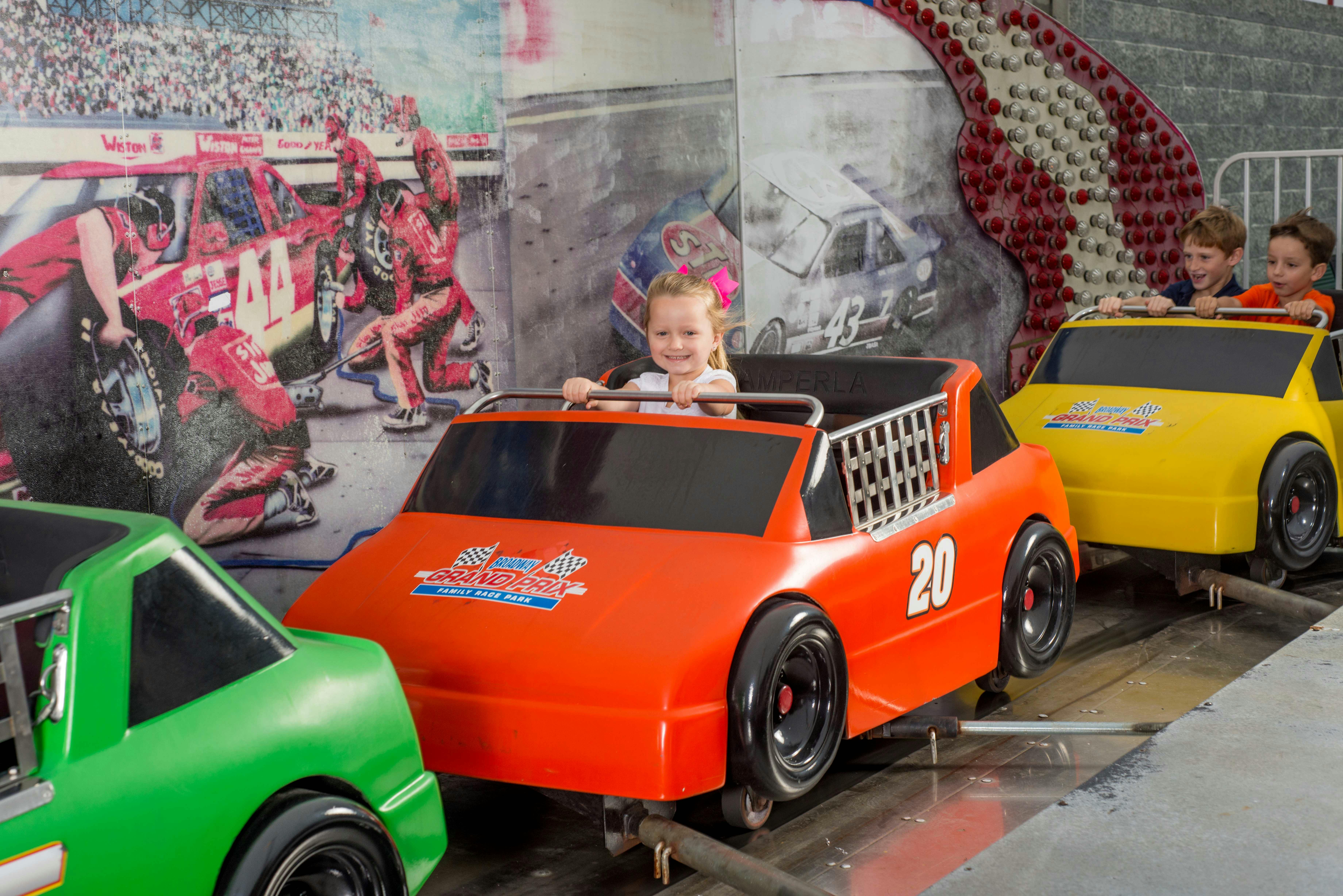 Young girl smiles while riding an orange car-themed ride with the number 20. Racing-themed backdrop and other colorful cars are seen.