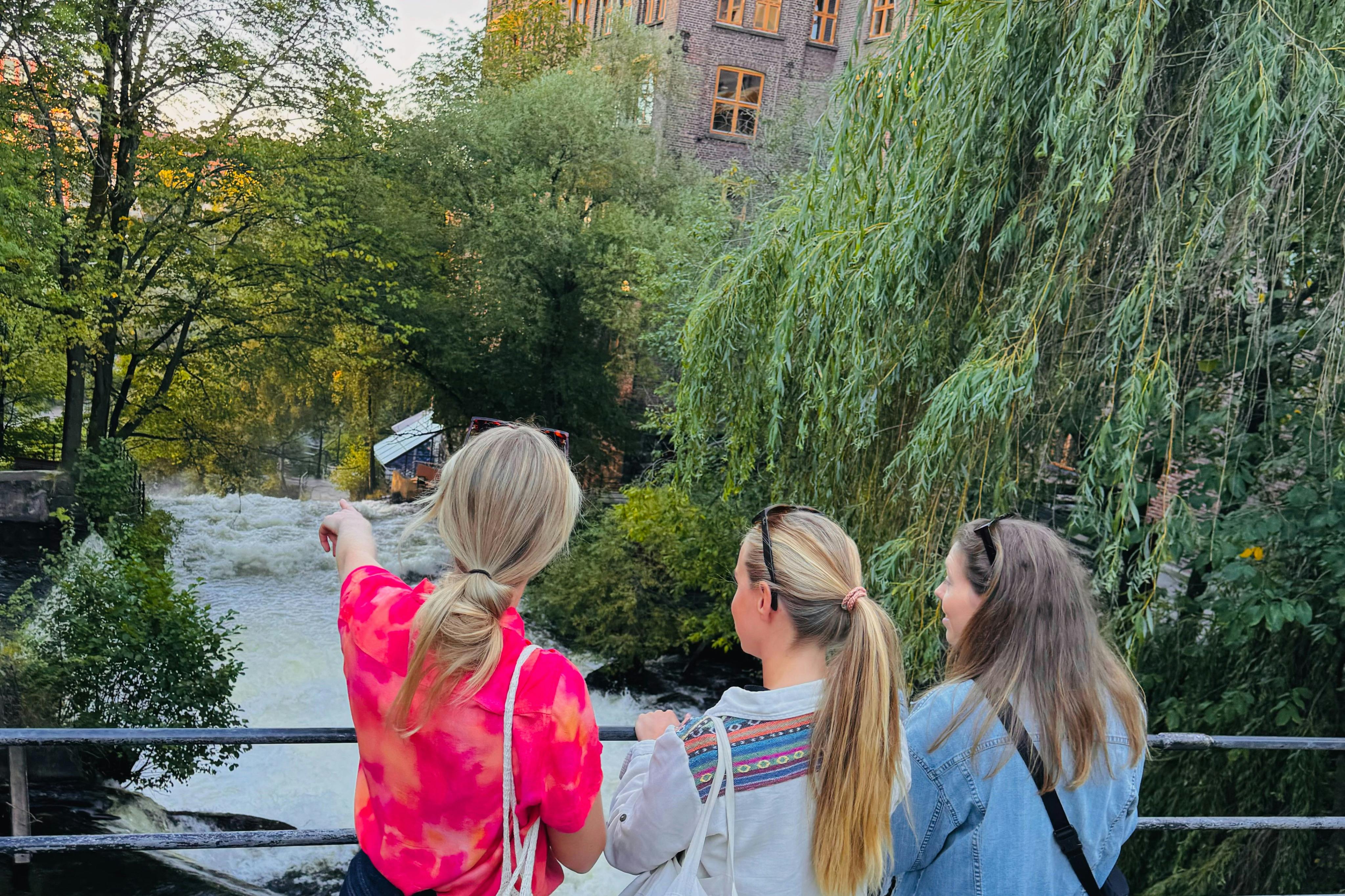 Three people with long hair stand on a bridge, facing away, with a large tree and a tall brick building in the background.