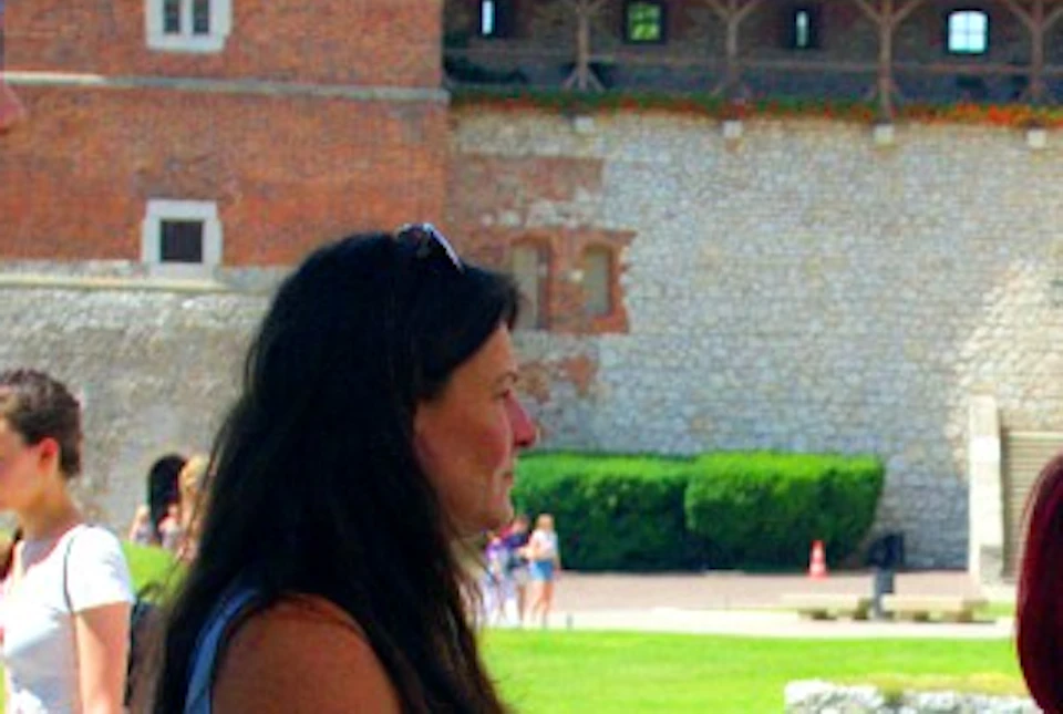 A group of people stands outdoors in casual attire, some looking at a historic building with a red roof in the background.