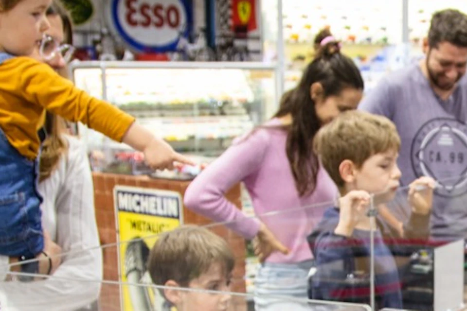Children and adults watch a model race car track in a toy store, with various toy cars on display.
