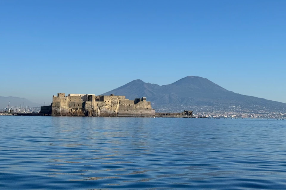 A historic stone castle on a coastal waterway with a mountain in the background under a clear blue sky.