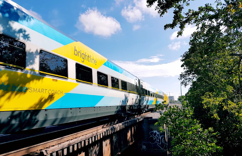A Brightline train crosses a bridge under a blue sky with scattered clouds, surrounded by greenery.