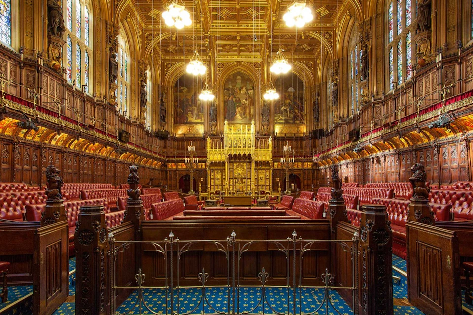 Ornate chamber with high ceilings, chandeliers, red upholstered benches, intricate woodwork, and large stained glass windows.