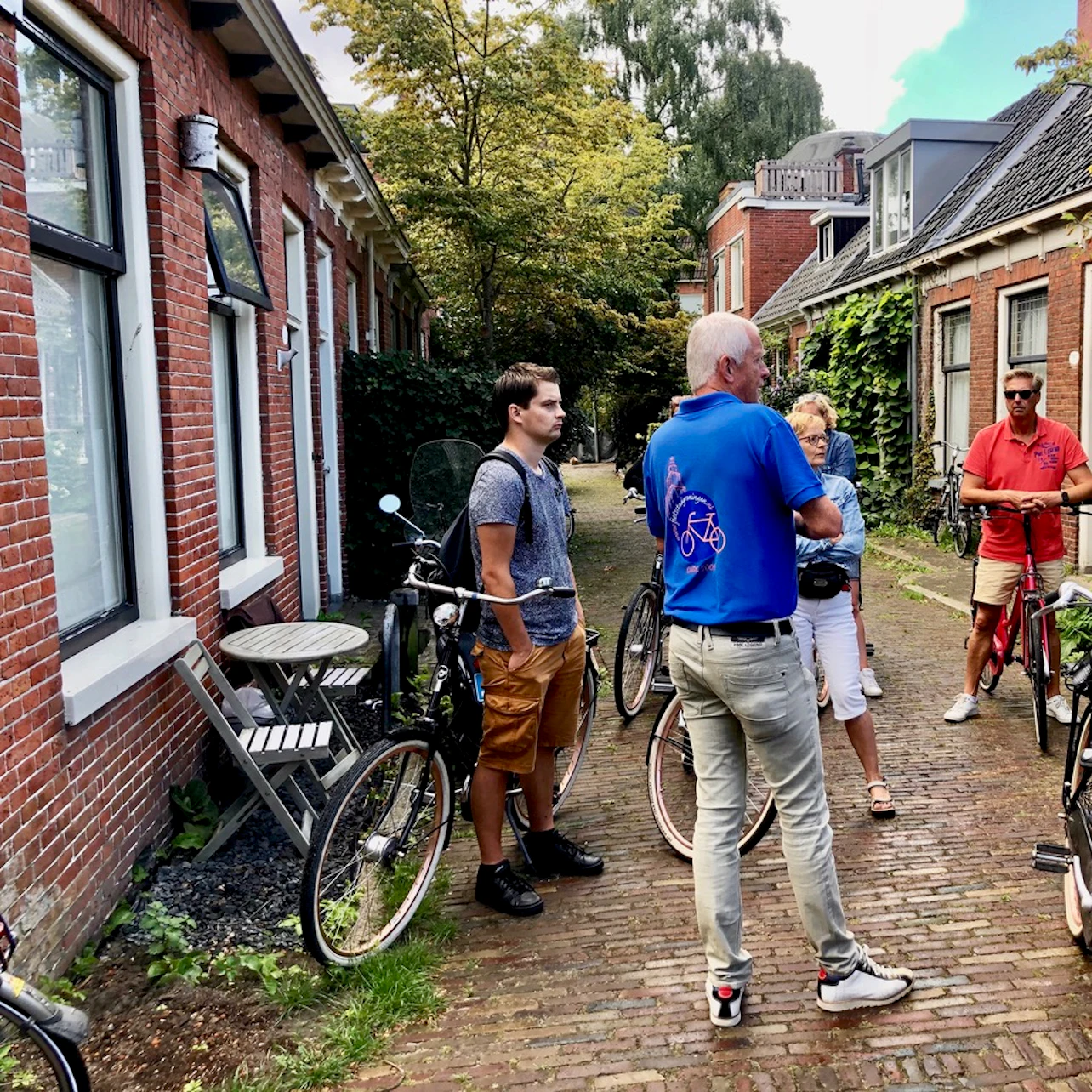 A group of people with bicycles stand and talk in a narrow, cobblestone alley between brick buildings with greenery.