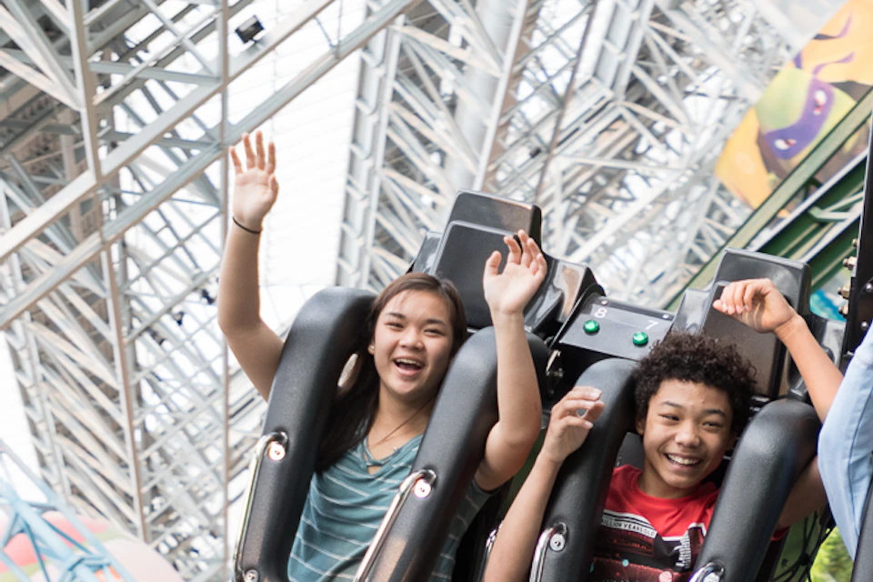 People on an indoor amusement park ride with raised hands, smiling and excited, surrounded by greenery and a steel frame structure.