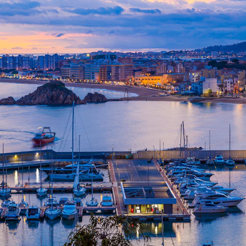 Marina med dockade båtar i skymningen, upplysta av gatubelysning. I bakgrunden en kuststadsmiljö med flerbostadshus och en liten ö.