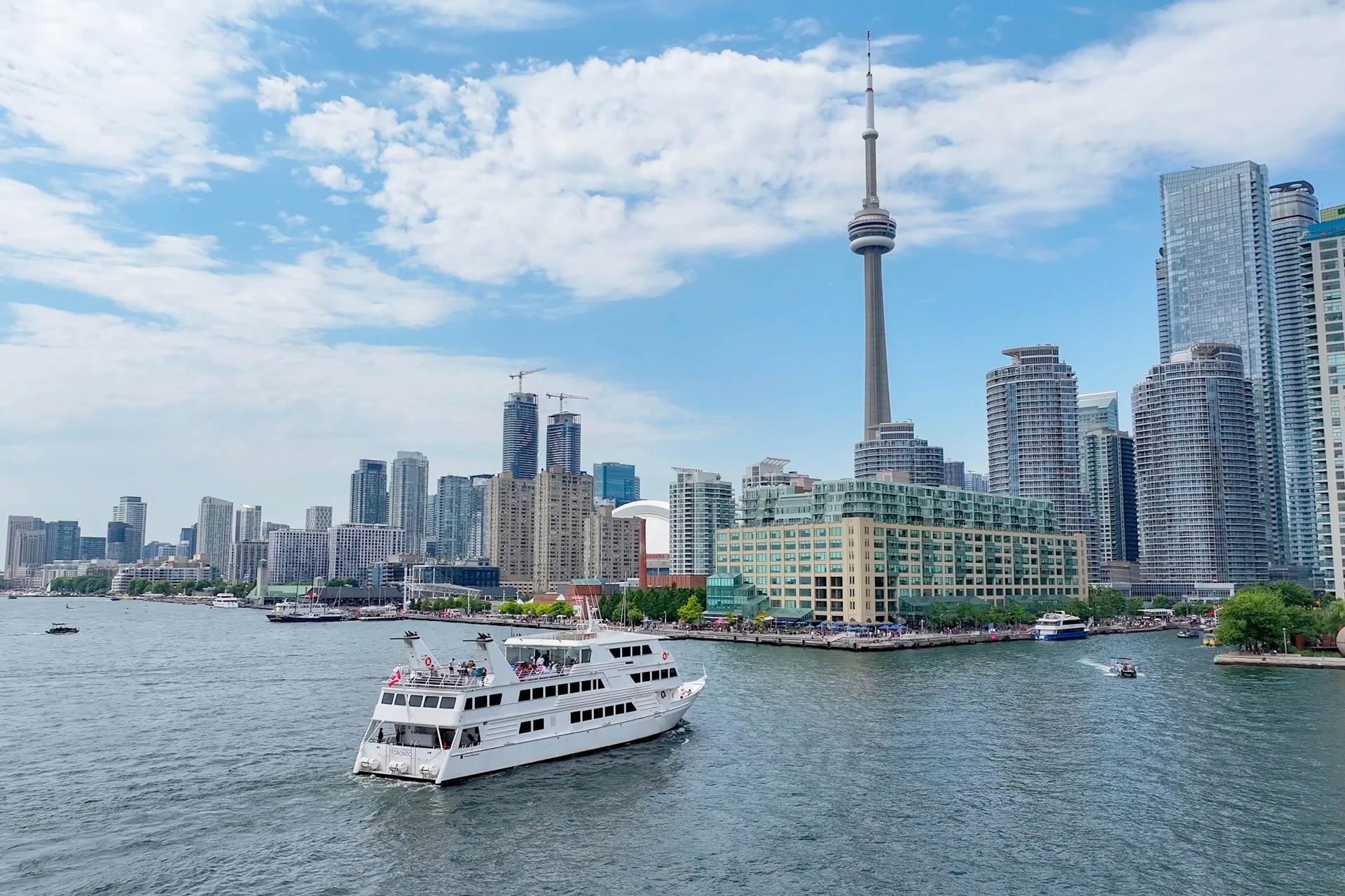 A white boat sailing on water with a city skyline and a tall tower in the background under a partly cloudy sky.