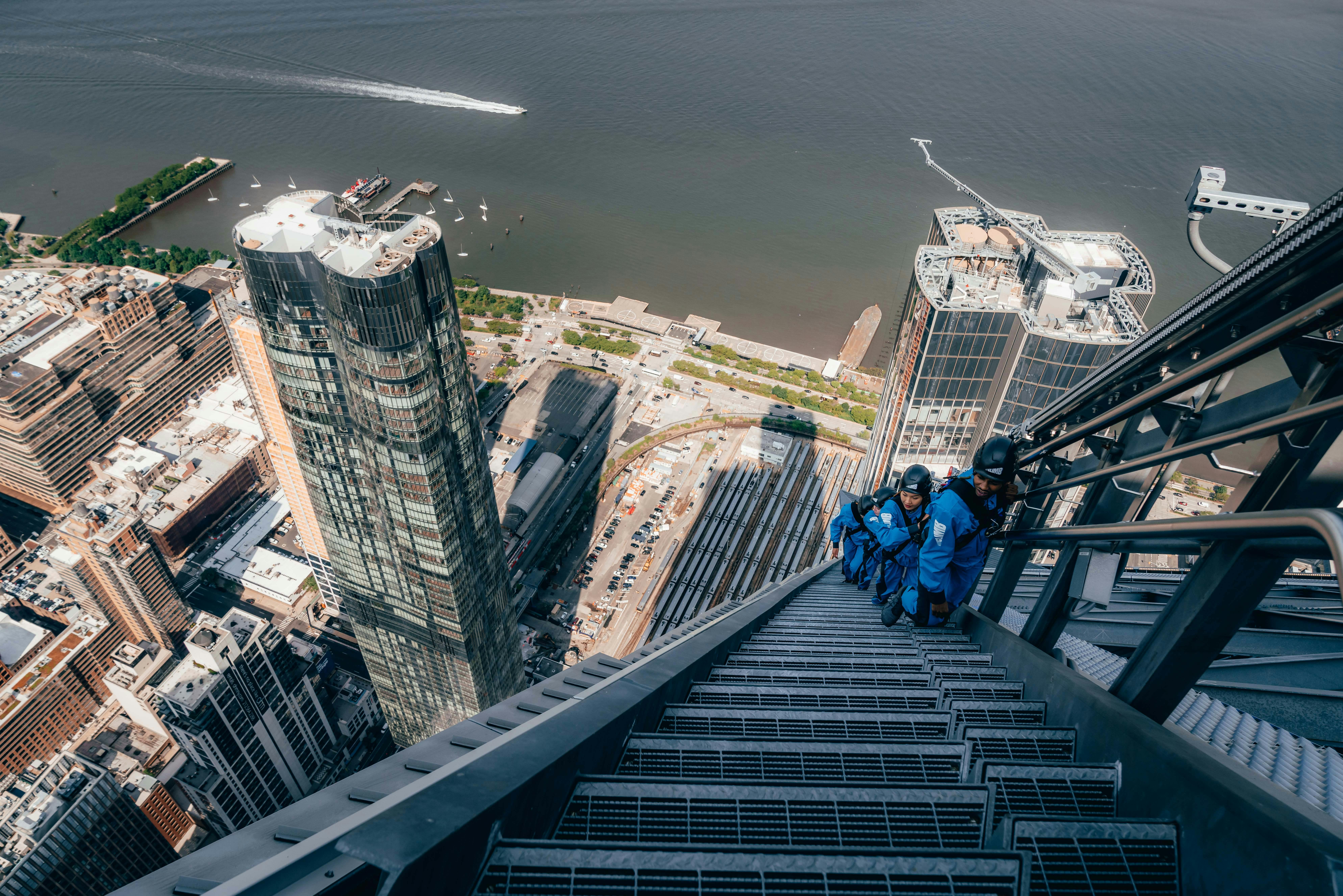 身穿蓝色攀岩装备的人们爬上摩天大楼一侧的楼梯，高耸于城市景观之上，后面有一条河流。