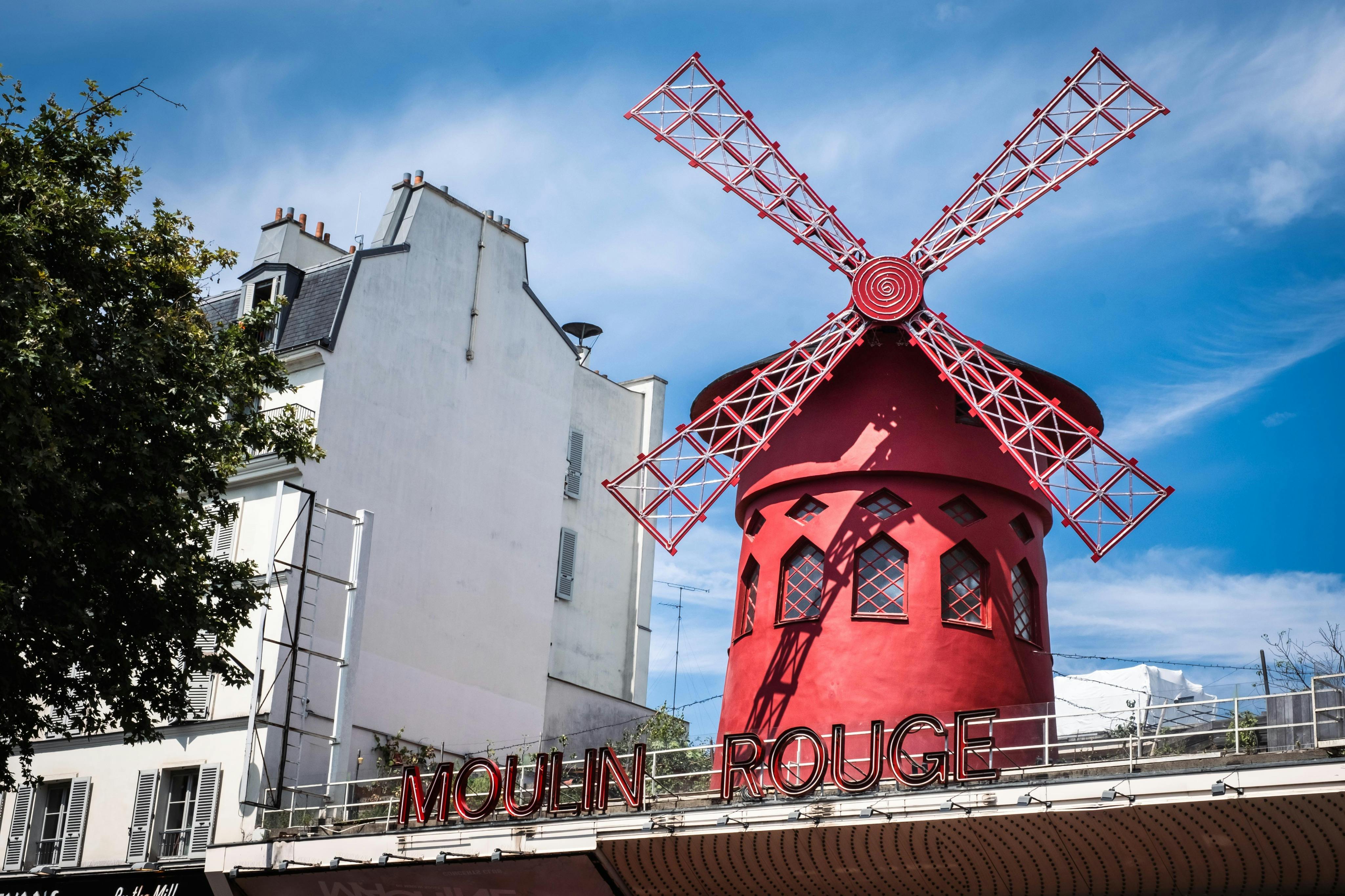 The Moulin Rouge building with its iconic red windmill against a blue sky, surrounded by other buildings.