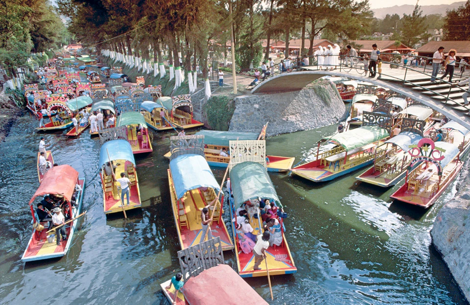 Colorful boats with canopies crowded in a canal under a stone bridge, surrounded by trees and people on the bridge and boats.